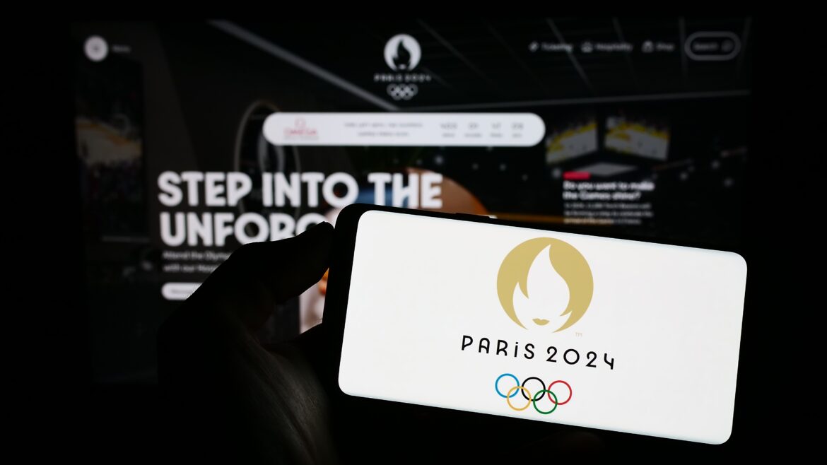 Person holding mobile phone with logo of the 2024 Summer Olympics in Paris on screen in front of web page. Focus on phone display. Unmodified photo.