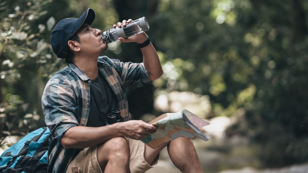 Young hiker taking a break for drinking water from the bottle to stay hydrated during hiking and holding a map in the other hand at the same time