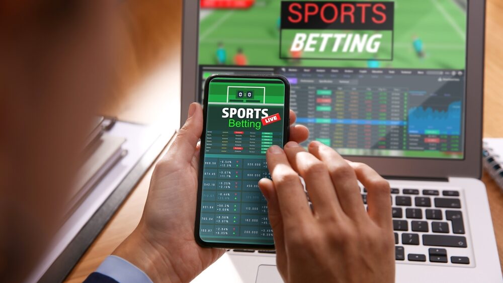 Man betting on sports using smartphone and laptop at table, closeup. Bookmaker websites on displays