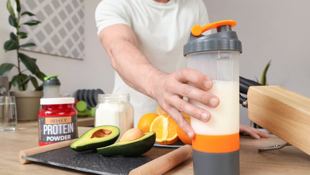 close up of muscular man picking up the shaker with pre workout mixture in it and avocado, orange and protein powder placed around it